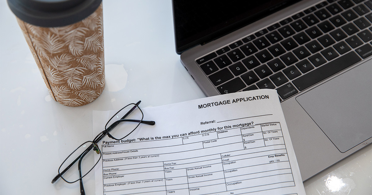 A coffee cup sits next to a laptop. There is also a pair of glasses and a mortgage application.
