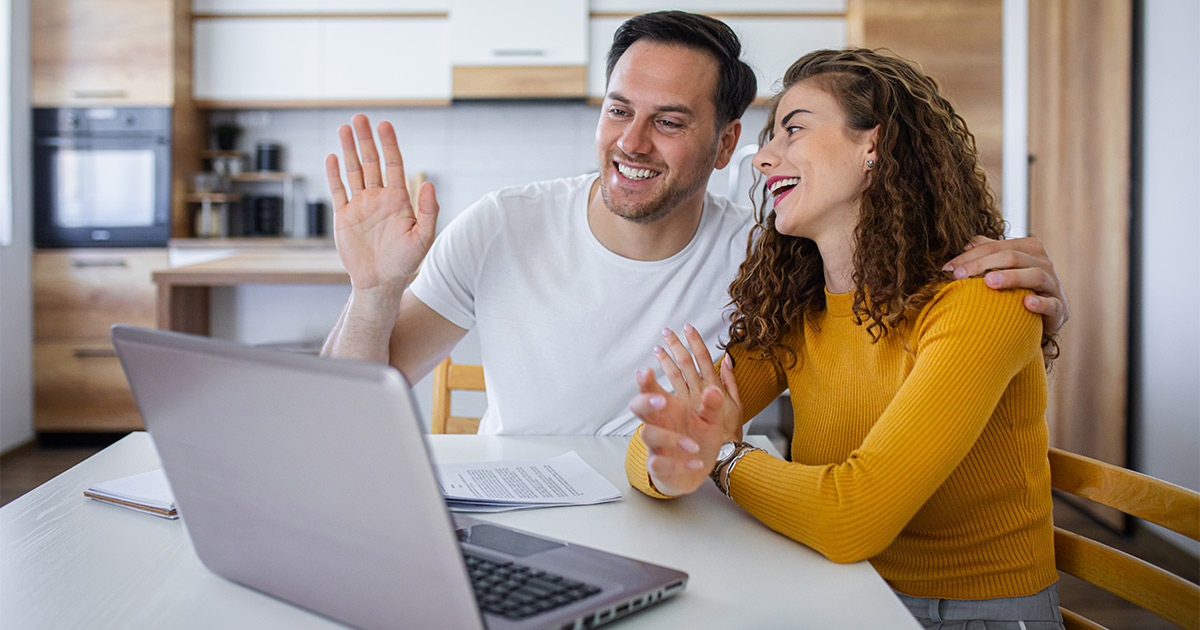 A young couple meets with their mortgage banker over a video call on their laptop computer. They have their paperwork for their upcoming long distance move in front of them on the table. 